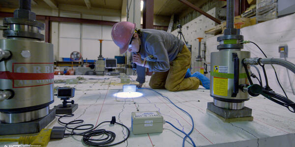 A researcher in a hard hat kneels on a concrete slab and uses a flashlight to examine it closely.
