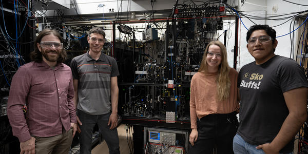Four researchers pose standing around a large piece of scientific equipment covered with wiring and electronics. 