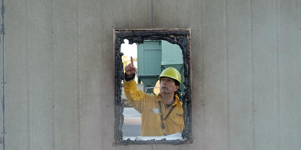 A man wearing a yellow hardhat and fire-resistant jacket points to burnt frame of a window.