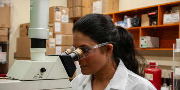 A woman wearing a lab coat and safety glasses stares into a large microscope.