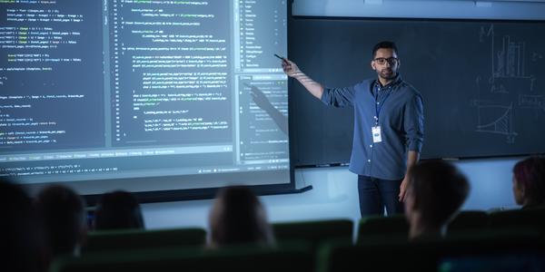 A young male teacher stands at the front of a darkened classroom, pointing at computer code on a large screen. 