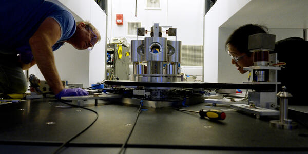 Wide shot shows two researchers on either side of a scientific device made of metal cylinders, peering closely at it. 