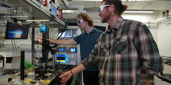 Two male researchers in casual clothes and protective eyewear stand in the lab, pointing at a screen hooked up to a laser science instrument. 