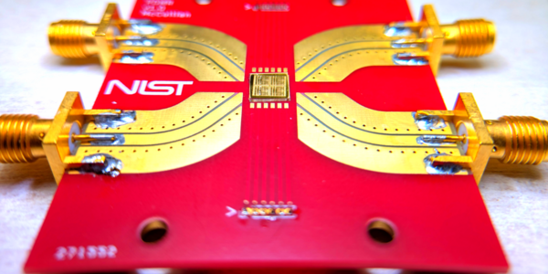 A bright-red rectangle with a NIST logo holds a gold-colored chip in the center with metal paths leading to connectors at the edge of the rectangle. 