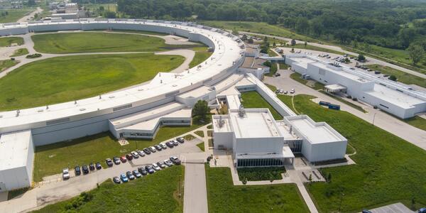An arial view shows part of the huge enclosed loop of an accelerator, with attached buildings and parking lots.