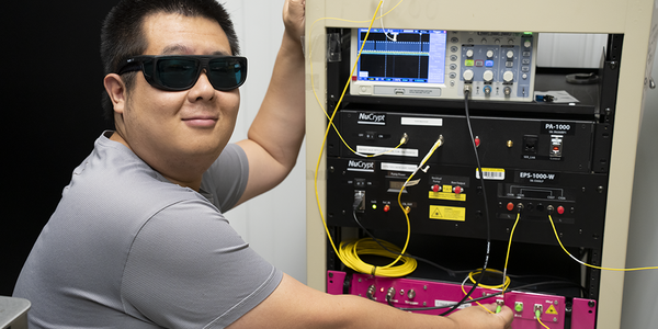 Yicheng Shi wears dark safety glasses as he poses next to a rack of electronics equipment.