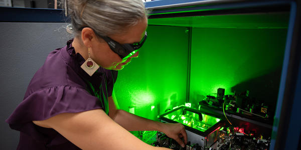 A researcher wearing safety glasses reaches into a box of circuitry and other equipment, which emits a green glow. 