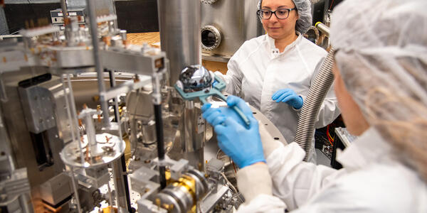 A researcher wearing a lab coat uses tongs to place a silvery sphere into a large scientific device as another researcher looks on. 