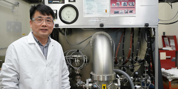 A researcher in a lab coat and safety glasses poses next to a large piece of lab equipment.