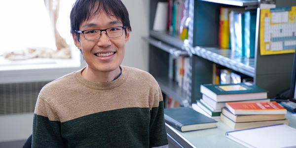 Leroy Jia poses smiling at his desk, with piles of books in the background. 