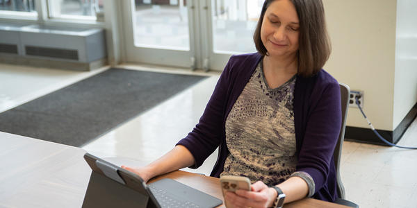Julie Haney sits at a table in the NIST library with a laptop, looking at a smartphone in her hand. 