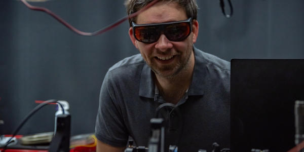 Krister Shalm, wearing safety glasses, smiles over a lab table covered with electronics equipment.