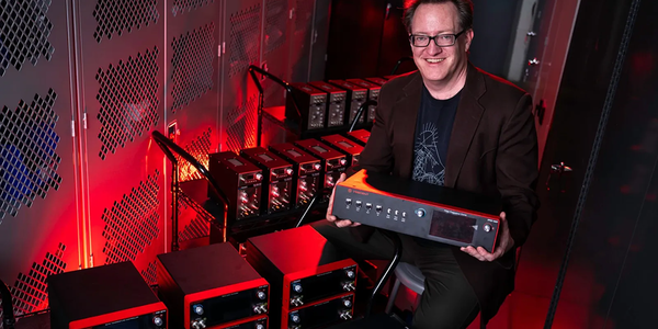 Scott Davis poses smiling and holding a frequency comb console. Other similar electronic products are stacked on carts around him. 
