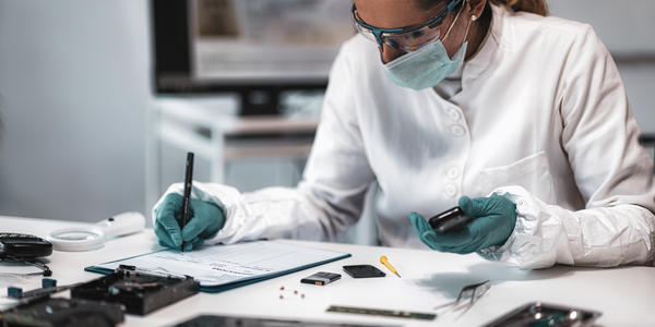A researcher wearing a mask, safety glasses and gloves sits at a desk, looking at electronic devices and writing on a clipboard. 