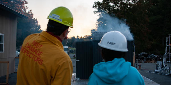 Two NIST researchers, wearing hardhats and protective jackets, watch a small shed burn as part of a setudy.