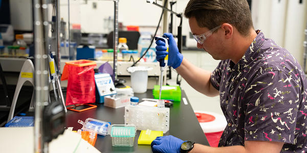 Researcher in safety glasses and gloves sits at a lab bench working with small test tubes.