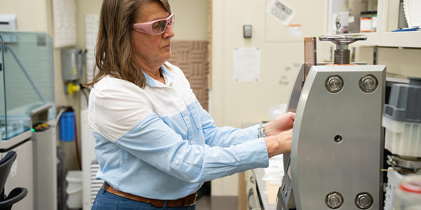A researcher wearing safety glasses stands in the lab, reaching into a metal device on the lab table. 