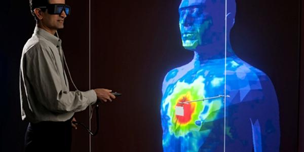 A researcher in safety glasses stands holding a remote control near a large blue model of a human head and torso with red marks on the chest. 