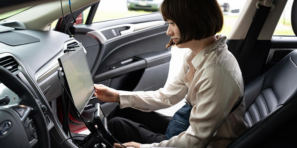 A woman sits in the passenger side of a parked car, looking at a large monitor attached to the dashboard. 