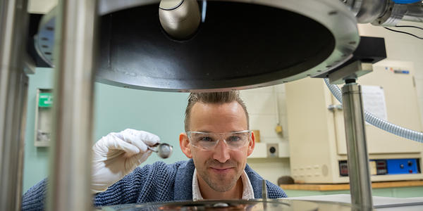 Andrew Iams wears safety glasses as his face is framed in an opening between two horizontal pieces of equipment in the lab. 