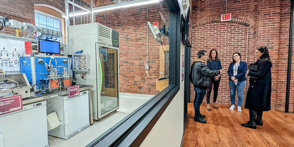 Four people stand talking in a converted factory space near a large interior window through which a complex, high-tech manufacturing line is visible. 