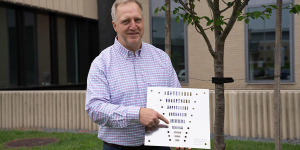 Jack Glover poses outside on the NIST campus, holding a flat square object with holes of varying sizes. 