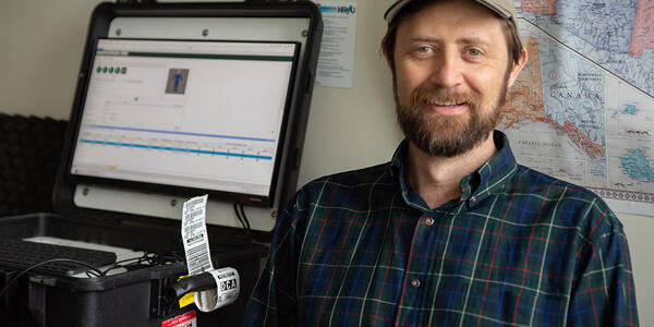 Gary Howarth poses for a photo in his office, wearing a NIST baseball cap, with a ruggedized laptop on the counter next to him and a map on the wall behind him.. 