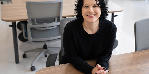 Kelly Sauerwein poses sitting at a table in the NIST library, leaning forward and smiling. 