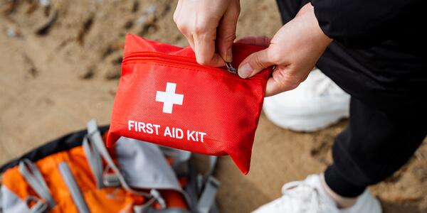 A person holds a red pouch labeled First Aid Kit as they stand over a travel bag on sandy ground.