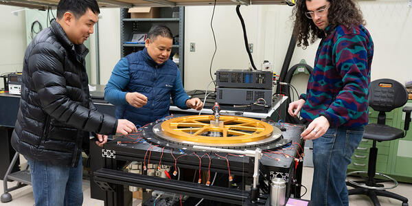 Three warmly dressed researchers stand around a scientific device with a large circular piece lying flat on top. 
