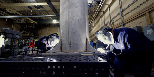 Two people wearing welding masks and other safety equipment bend over a table to work on a large metal cylinder.