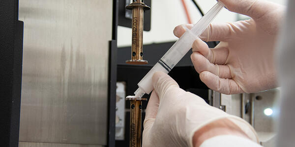 Researcher holds a small white tube filled with wet ceramics and points it toward lab equipment.