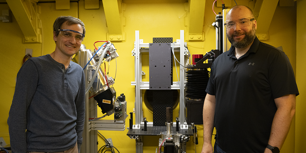 Cyrus Daugherty and Jake LaManna, wearing safety glasses, pose on either side of an imaging device holding a replica scroll.