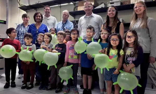 Adults standing in the back row. Kids with blown up green gloves stand in the front row.