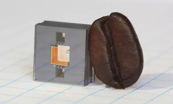Square gray cube with two square holes in it and a gold square in between the holes, and a dark brown coffee bean leaning against the cube.