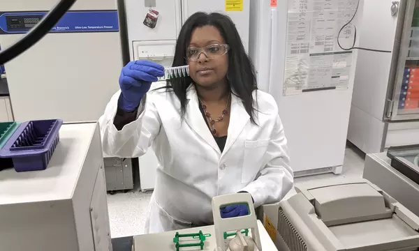 young woman in a lab coat looks at vials with a small amount of green liquid in them before putting them into a machine