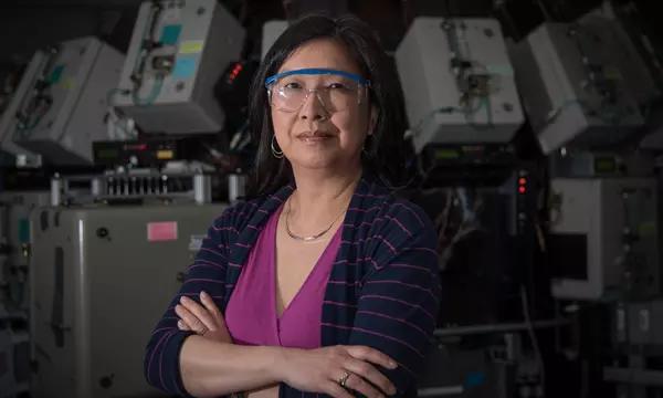 Joannie Chin stands with arms crossed, wearing safety goggles, in front of scientific equipment. 