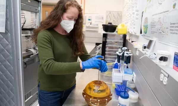 A woman wearing a mask stands over a yellow sphere at a lab bench. 