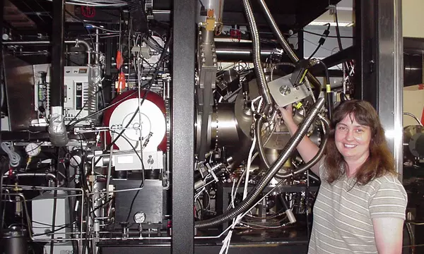 A woman stands smiling in front of a large device with many metal parts and wires.