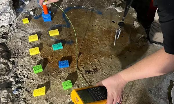 A researcher with a handheld device performs tests with small pieces of colored material lying on a large piece of concrete.