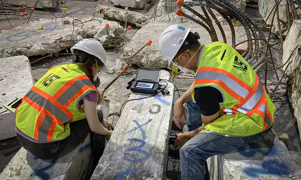 Two people wearing safety gear sit on either side of a concrete column lying on the floor, performing tests on it.