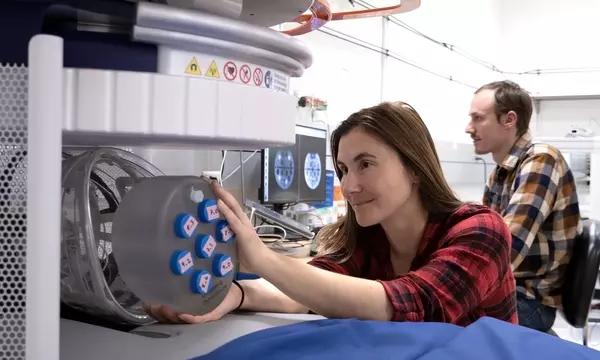 A woman leans over an MRI machine to reach a set of samples in a round holder, while a man sits behind her at a computer looking at MRI images. 