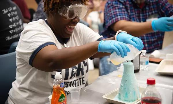 Kandice Taylor, in safety glasses, laughs as she pours liquids into a glass flask that is overflowing with foam.