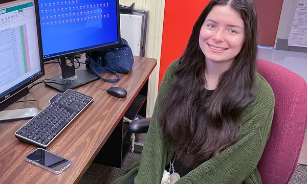 Isabelle Rivera poses smiling at a computer with two monitors, one showing a spreadsheet and the other multiple file icons. 
