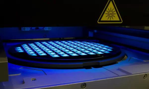 Photograph depicting IC manufacturing showing glowing squares on a round wafer, with a yellow triangular warning indicator labeled UV.