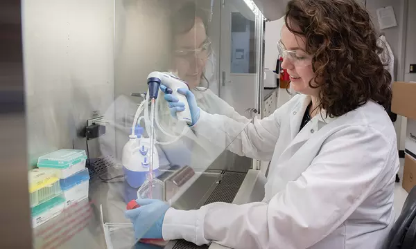 Jerilyn Izac sits at a lab bench with her hands inside a plastic shield, using a device to draw liquid from a plastic container into a tube. 