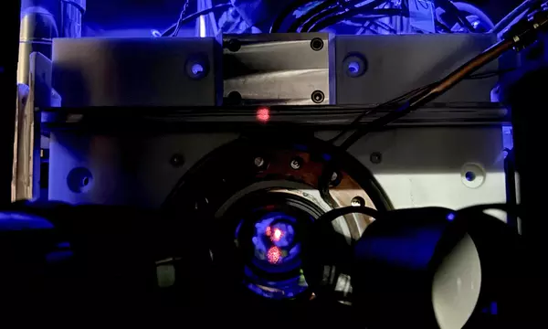 Close-up of a scientific instrument with wires and a circular metallic component, illuminated by blue light, featuring a small red dot at the center.