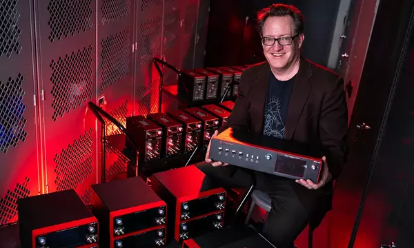 Scott Davis poses smiling and holding a frequency comb console. Other similar electronic products are stacked on carts around him. 