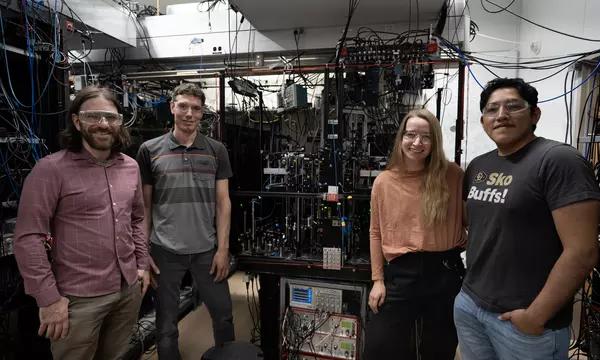 Four researchers pose standing around a large piece of scientific equipment covered with wiring and electronics. 