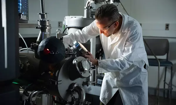 Andrew Iams wears a lab coat and safety glasses as he looks into a circular opening on a large piece of lab equipment. 